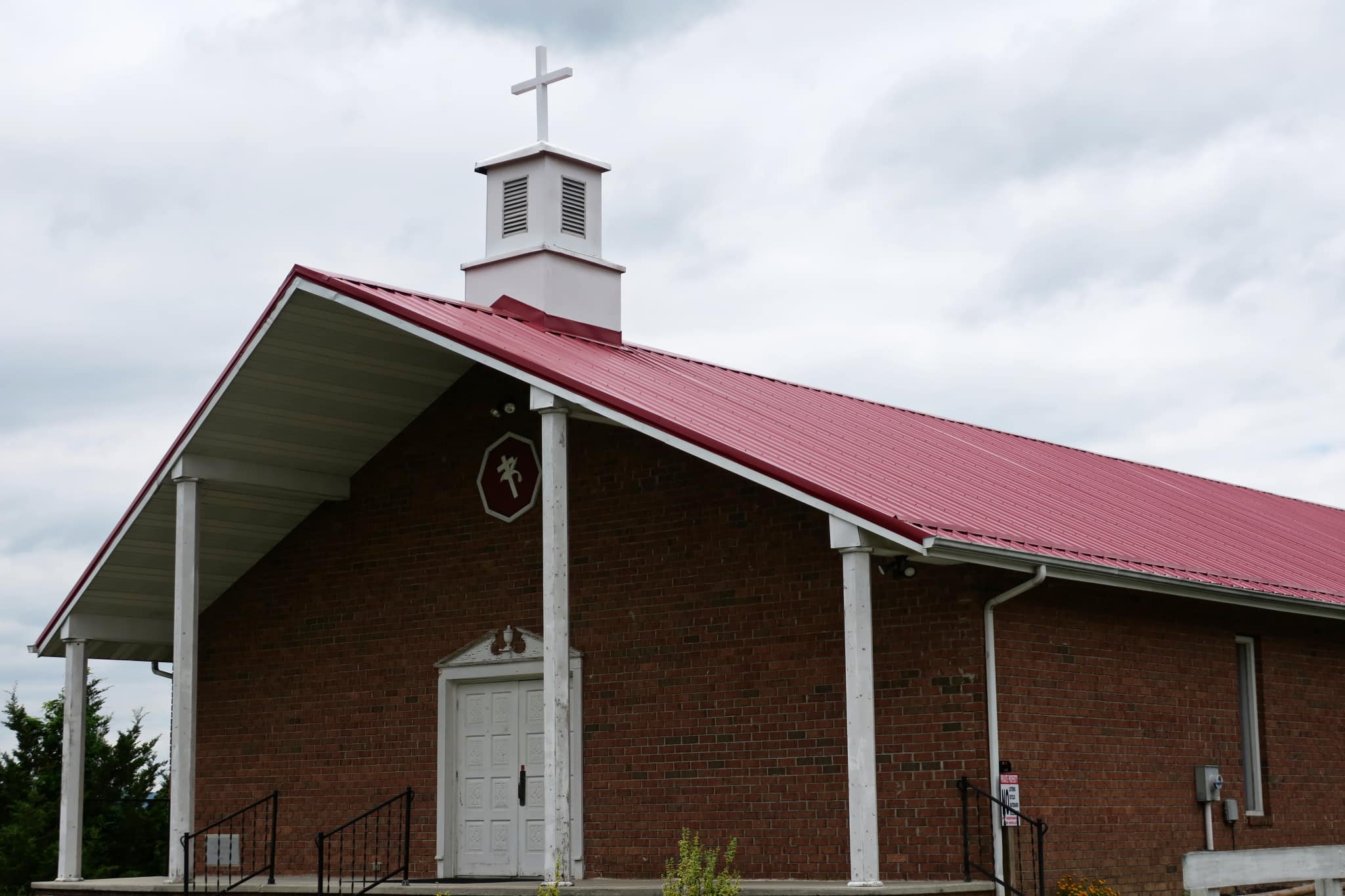 A brick church building with a red metal roof, white doors, and a cross on top under a cloudy sky.