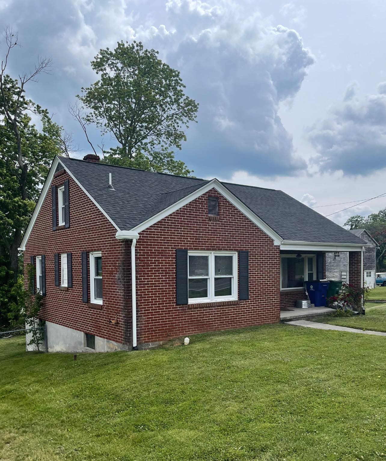 A red brick house with black shutters, white trim, and a gray shingle roof sits on a grassy lawn under a partly cloudy sky.