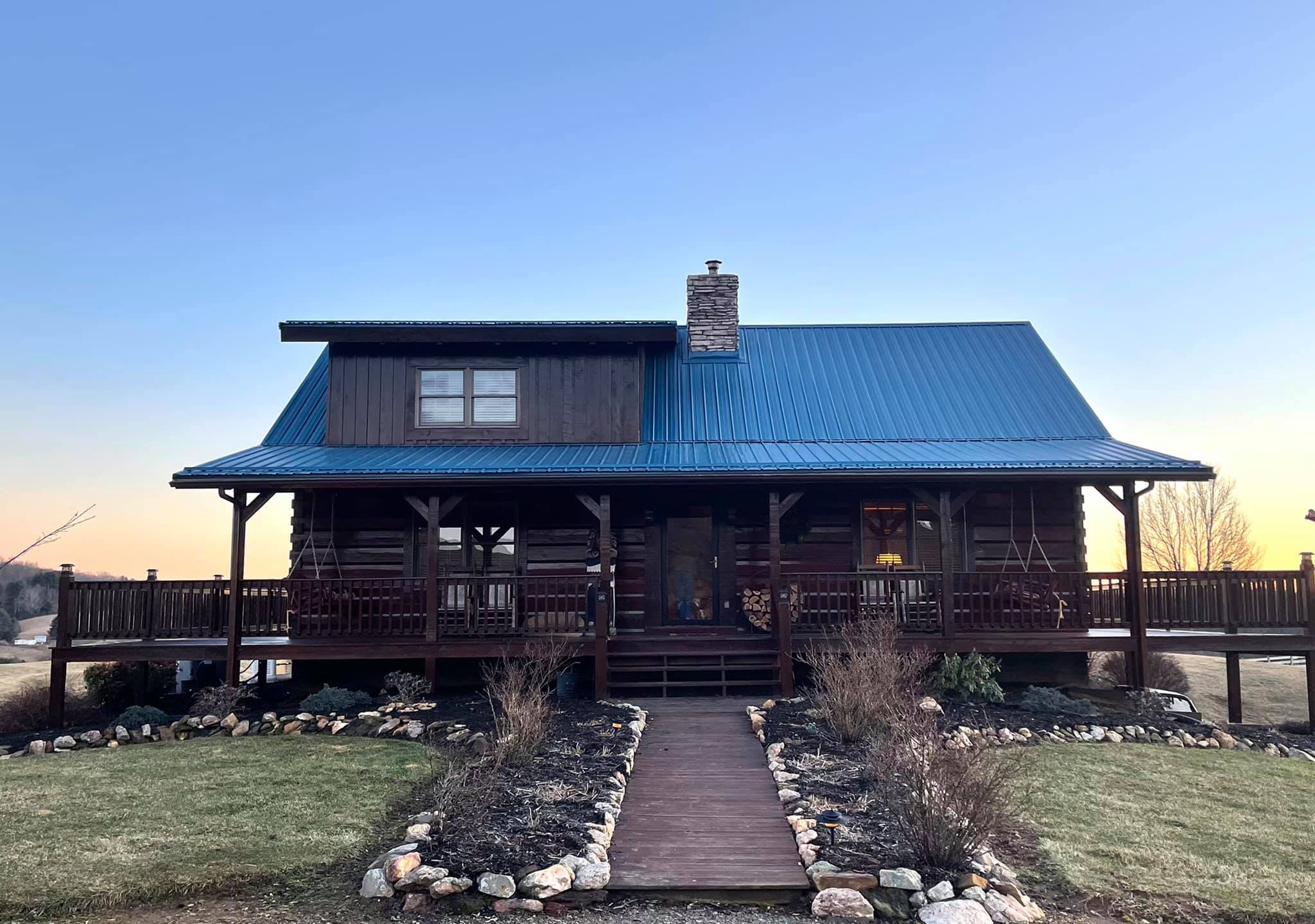 Log cabin with a blue metal roof, wraparound porch, and stone-lined walkway, surrounded by a grassy yard and landscaping, photographed at sunset.