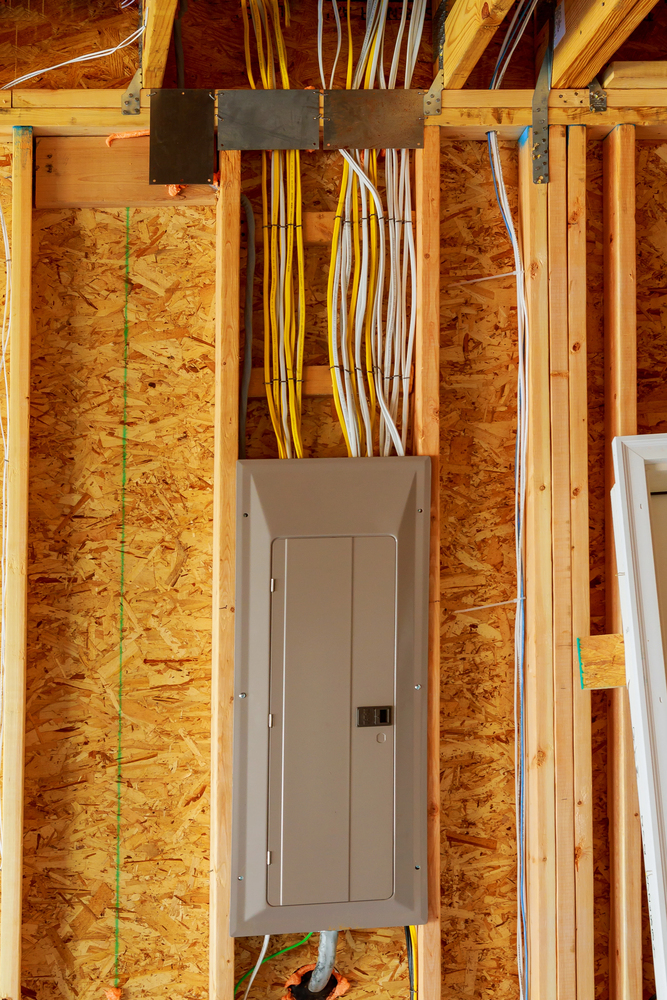 A gray electrical panel is mounted on an unfinished wooden wall with multiple yellow and white wires running above it.