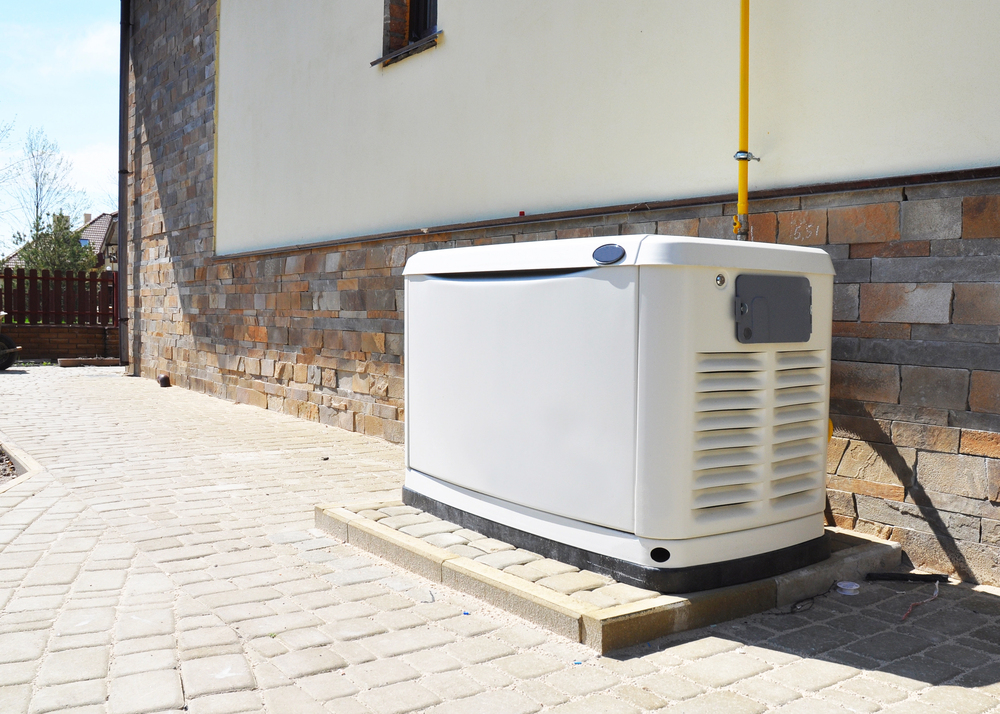 A standby generator installed on a paved area outside a house with a stone and stucco exterior wall.