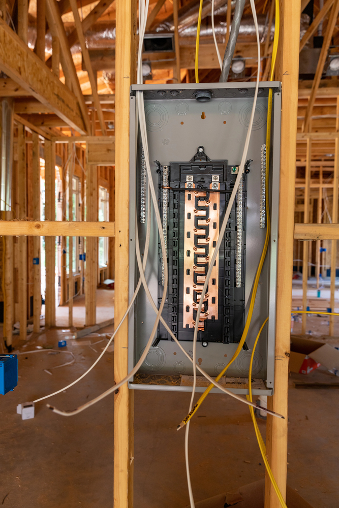 An electrical panel with exposed wiring is mounted on a wooden frame in a house under construction.