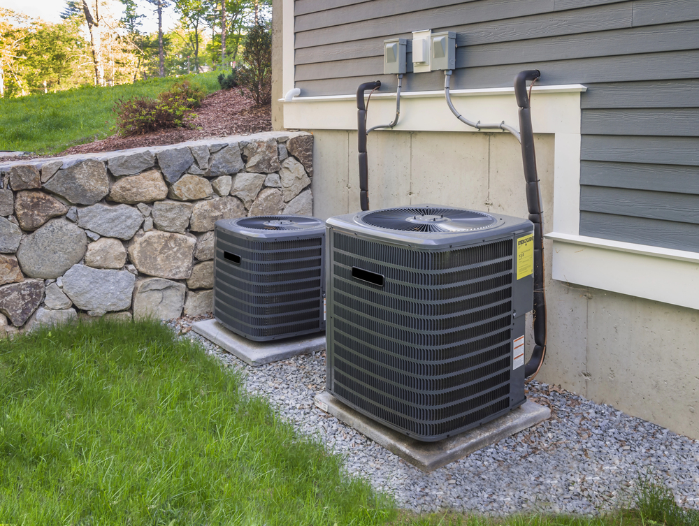 Two outdoor air conditioning units are installed on gravel pads next to a house exterior with stone retaining wall and electrical boxes visible.