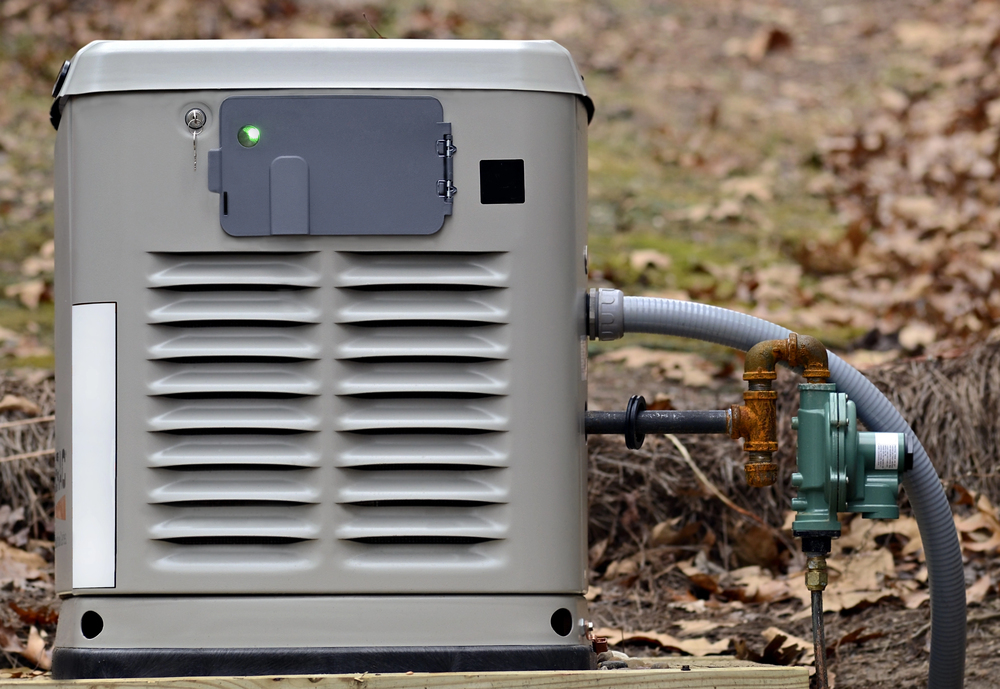 A standby home generator with a green indicator light is connected to a gas supply line outdoors, surrounded by dry leaves.