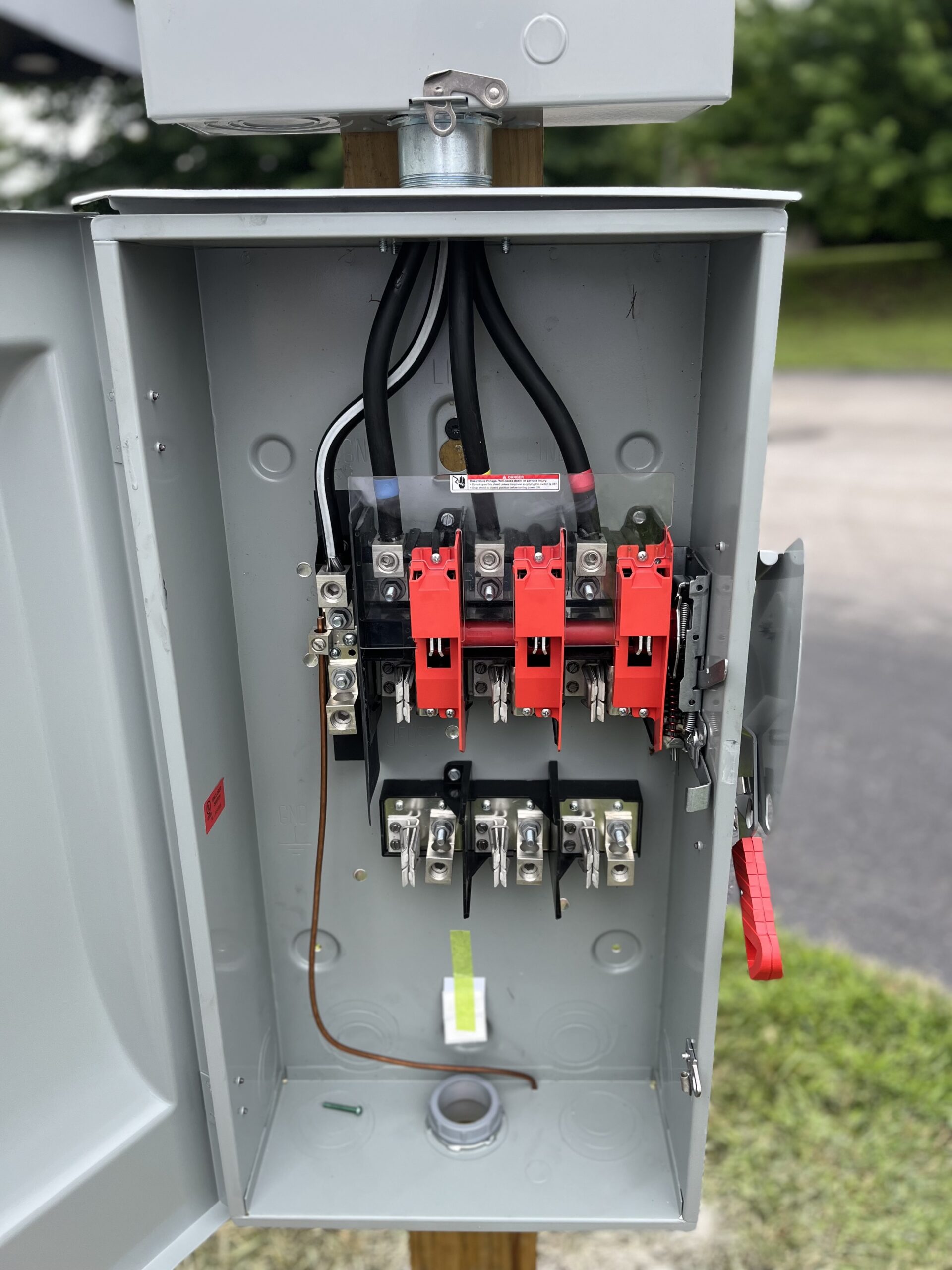An open electrical panel with visible wiring, switches, and fuses, mounted on a wooden post outdoors.
