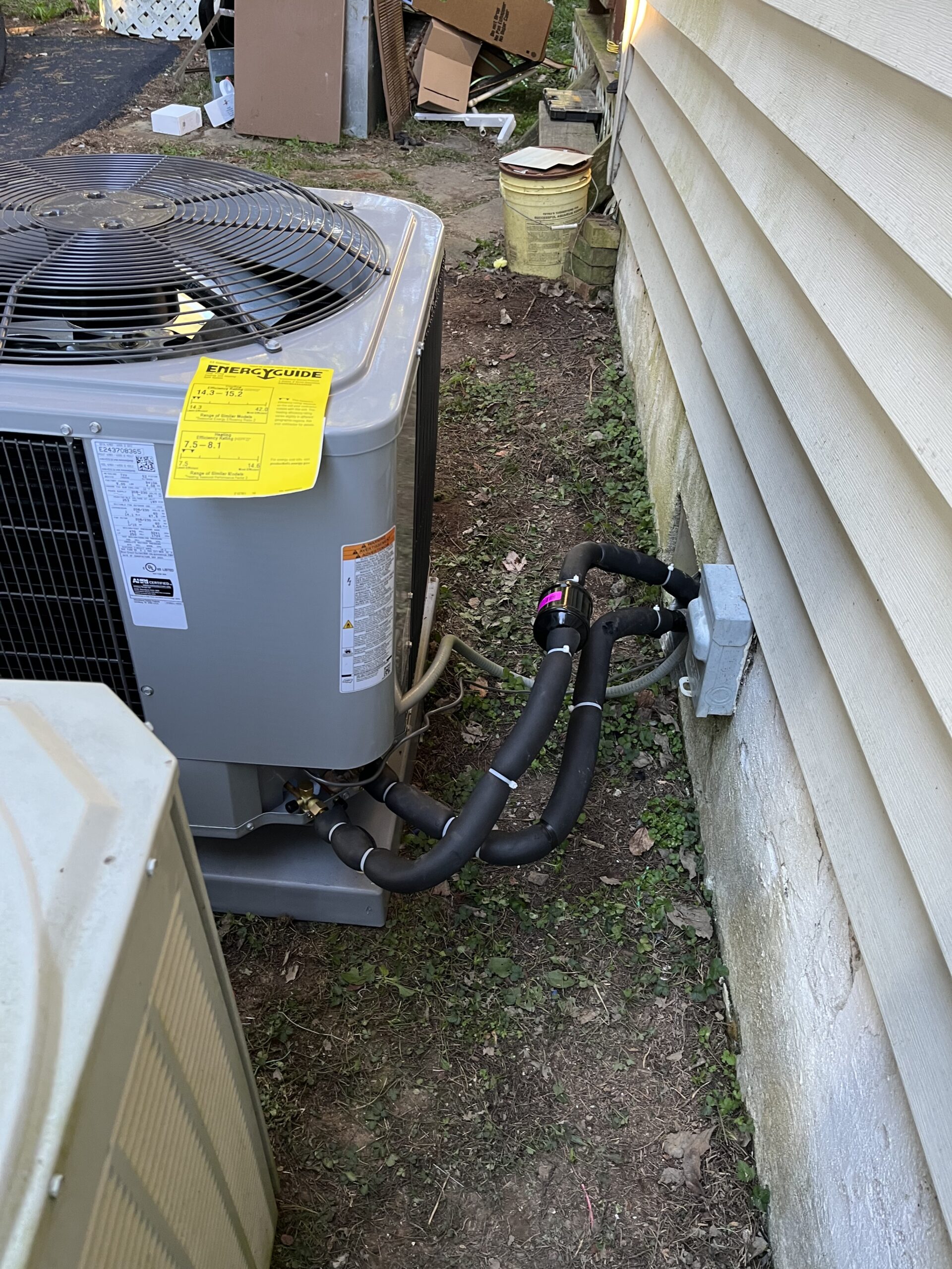 An outdoor air conditioning unit is installed beside a house, with insulated refrigerant lines connected to an external wall. An energy guide label is attached to the unit.