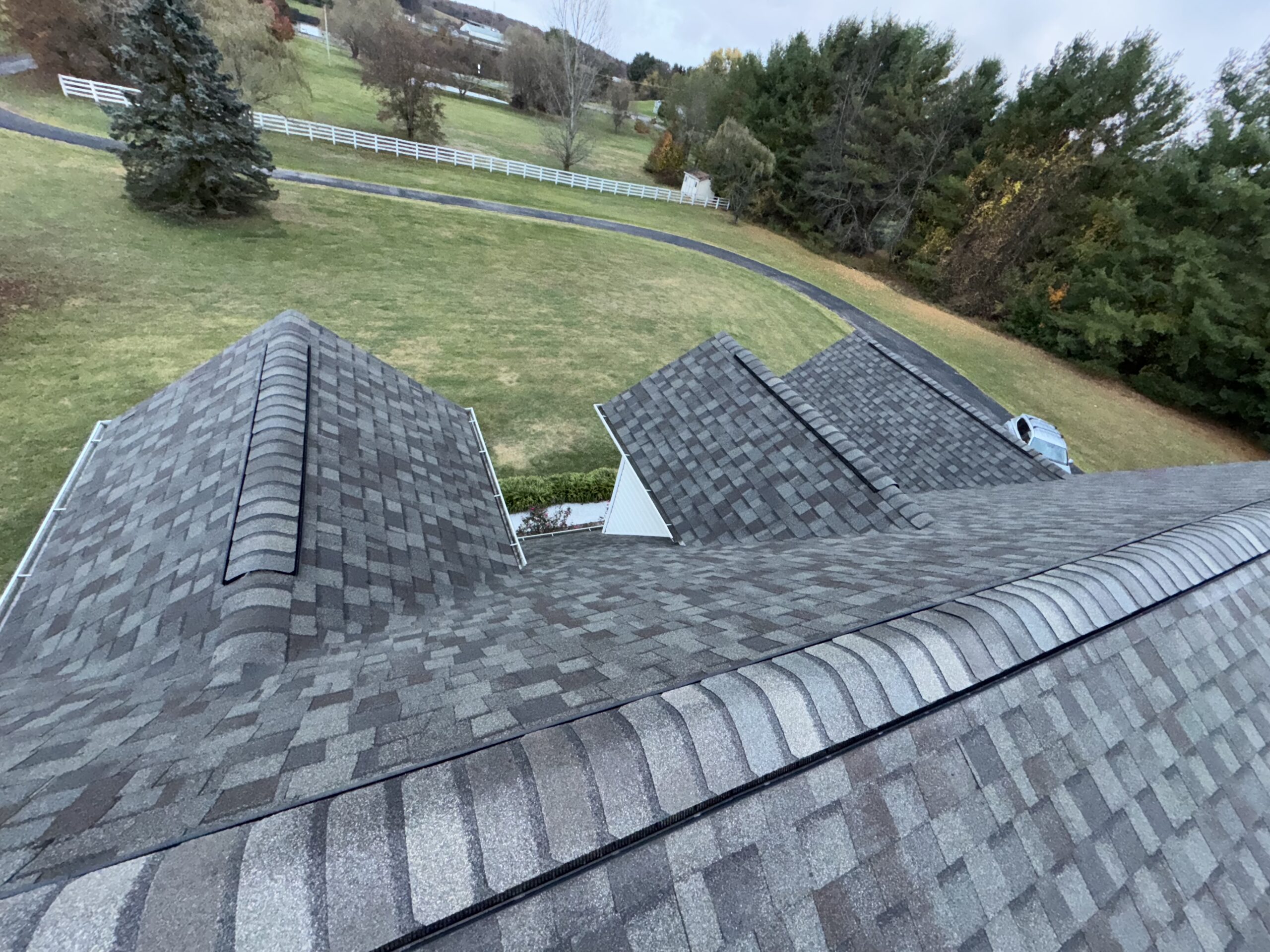 Aerial view of a house roof with gray asphalt shingles, surrounded by a large yard, trees, and a winding driveway.