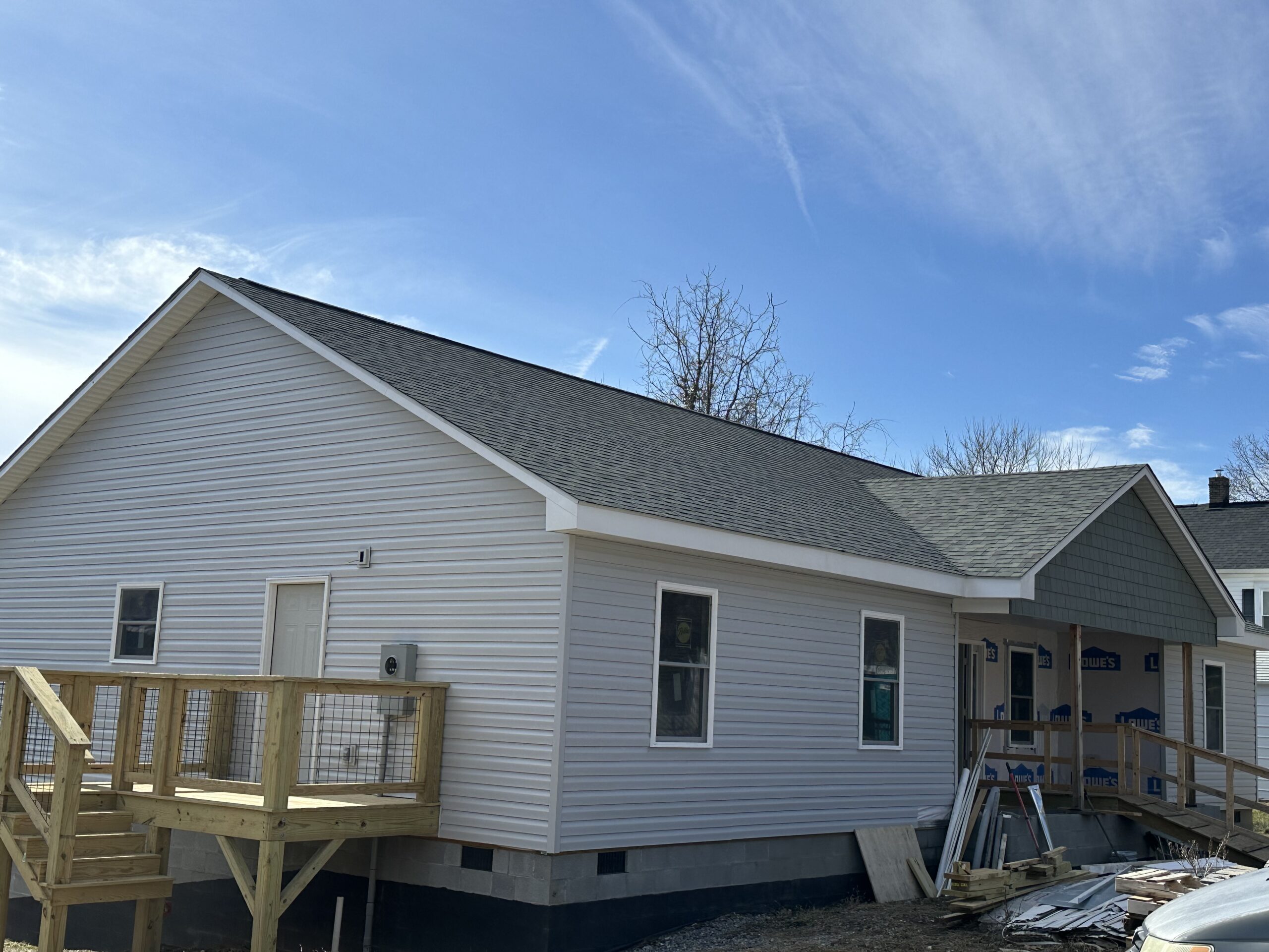 Single-story house with light-colored siding, gray shingled roof, wooden porch, and construction materials scattered nearby under a clear blue sky.